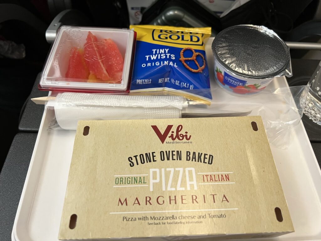 A box with the words "Pizza Margherita" on it, alongside a small cup of fruit, a bag of pretzels and a yogurt on the JAL tray table.