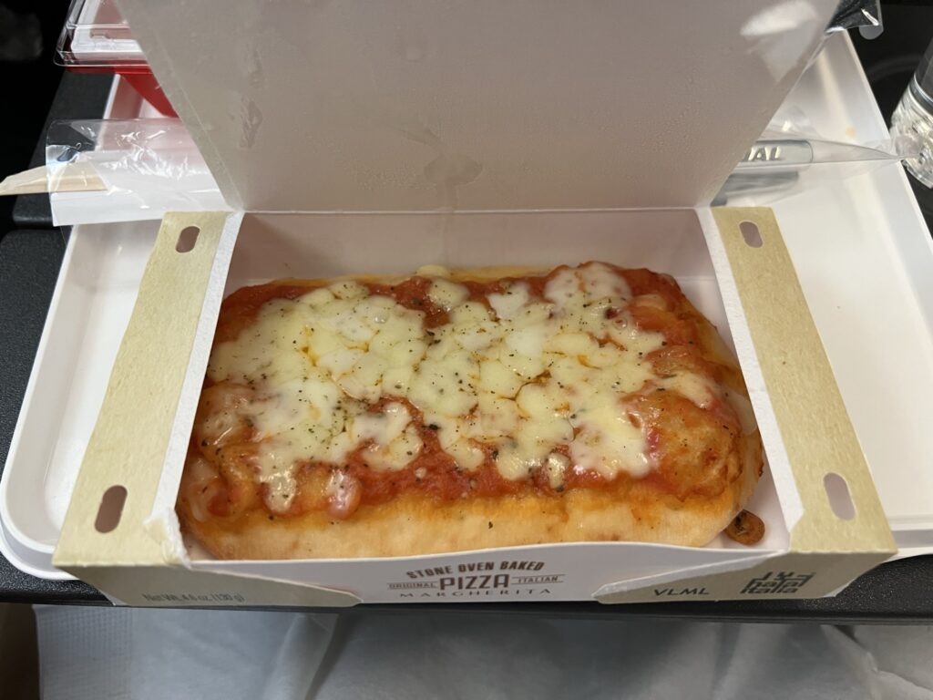 A slice of pizza sits in a cardboard box atop the JAL tray table.