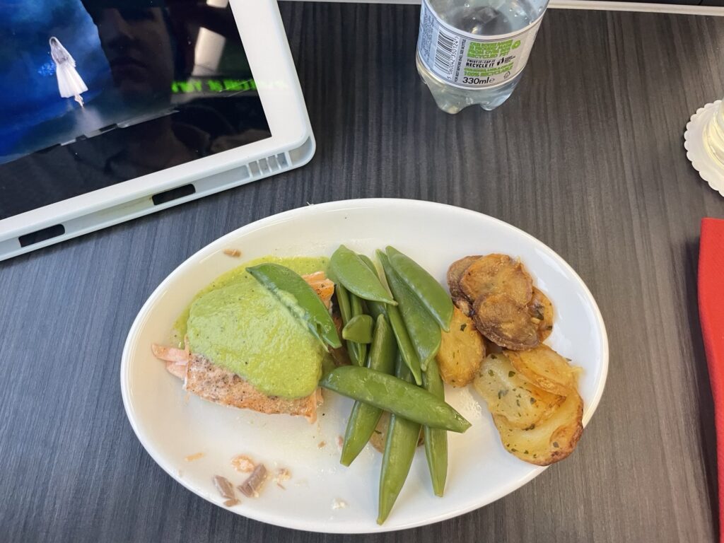 On board the train, a meal of potatoes, salmon and green peas sit on a table near a tablet that is playing a movie.