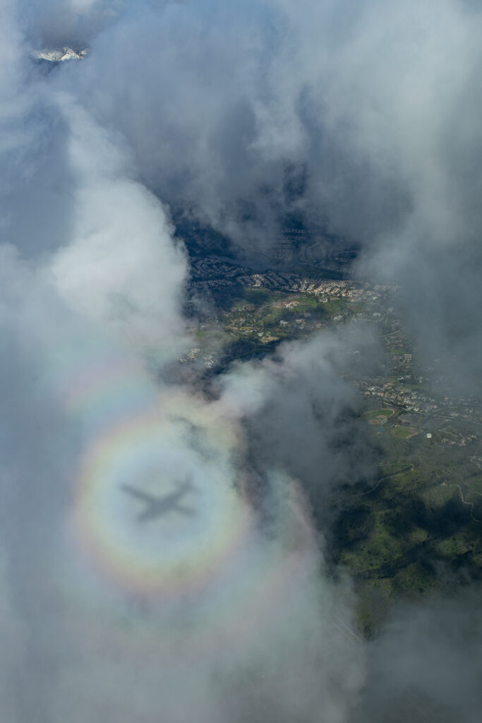 A reflection of the aircraft is visible in the clouds below, as the aircraft flies above beautiful scenery. 