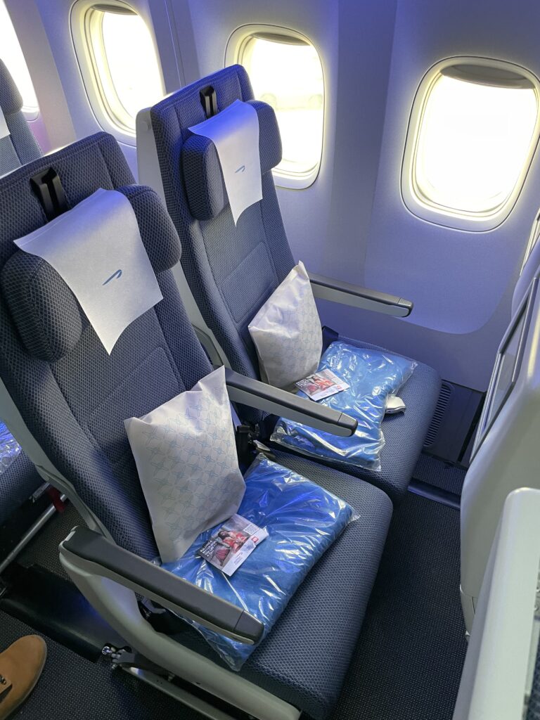 British Airways economy class seat with an amenity kit, blue blanket and white pillow on top of the seat bottom, waiting for the passenger.