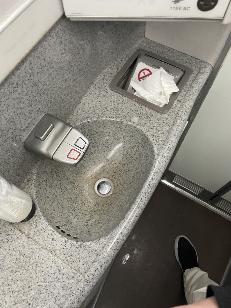 The sink in the British Airways 777 lavatory is speckled grey. The floor is already wet, and there is paper hanging out of the garbage bin.