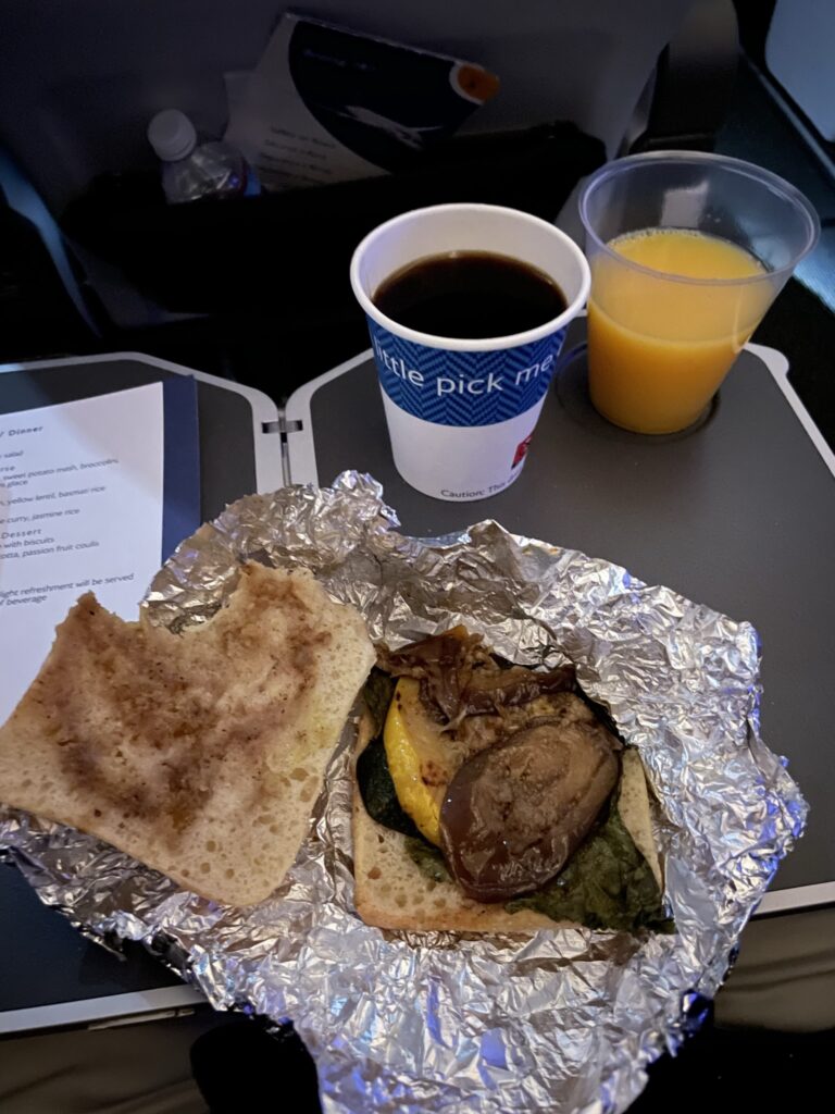 A horrible looking breakfast sandwich in foil and a coffee sit on the aircraft tray table. 