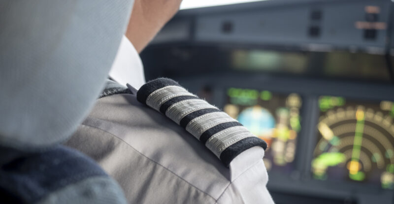 Over the shoulder shot of a captain flying a large transport aircraft. The four stripes on his uniform are in clear view, whilst avionics and displays are seen in front of him.