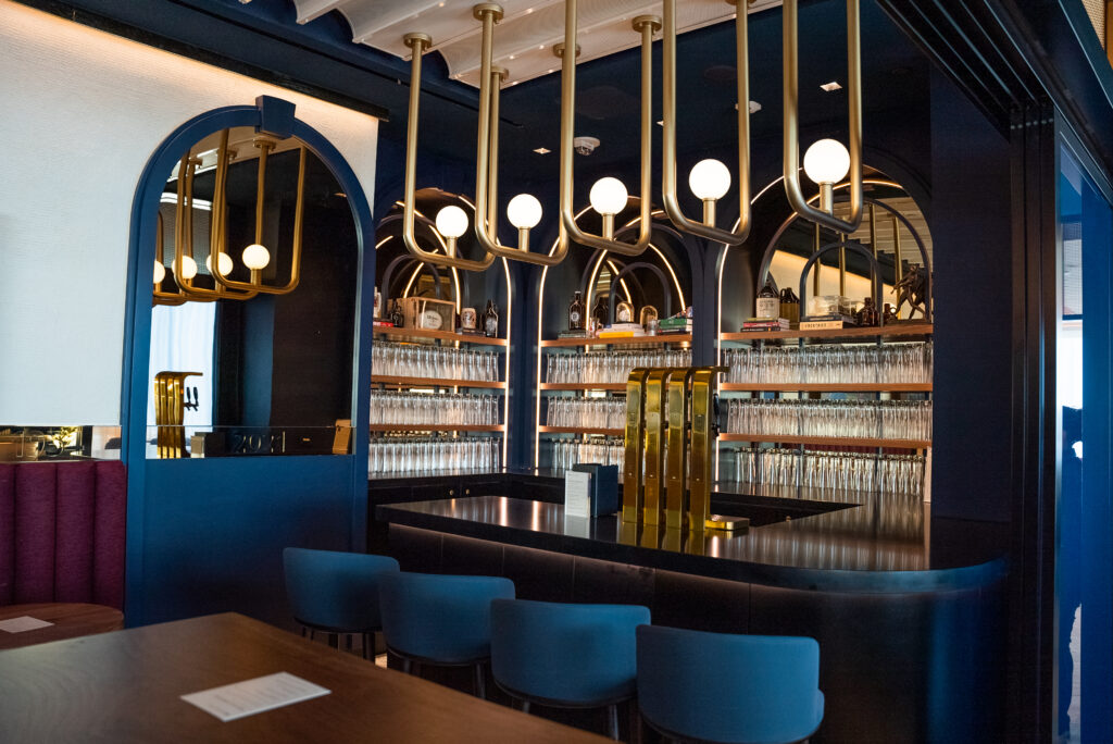 Gold lights hang above a bar ,with blue cushioned stools lined up at the bar as well.