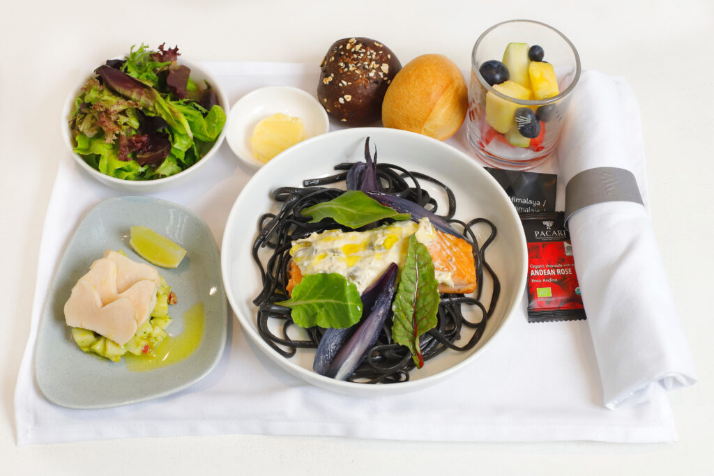 A tasty looking meal on board, including a large bowl of black noodles with various veggies, plus various side dishes, all elegantly laid out on nice dishes and a white cloth.