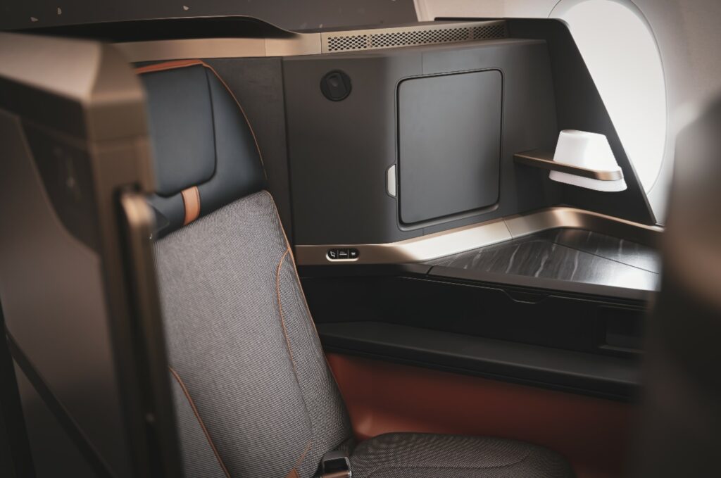 The Elements suite on a Starlux A350 seat. The seat is in grey, with a black headrest, and accented by copper and burnt orange. A halo like lip on the shroud angles inward for additional privacy.