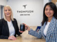 Two women execs at Thompson showcase the premium aircraft seatmaker's new logo on a coffee mug, whilst standing in front of the logo displayed on the white wall behind them.