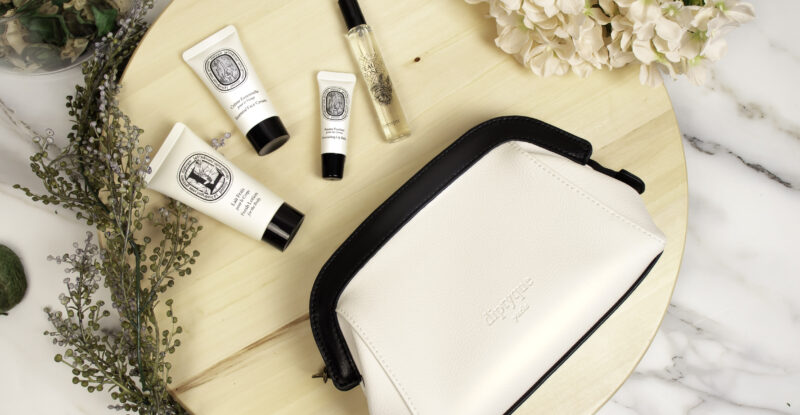 A sleek white amenity kit trimmed with black on a wood table, with various items displayed including lip balm and moisturizer.