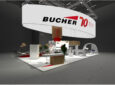 Bucher stand at AIX 2023 on the show floor. A large white circular sign is above the stand and says Bucher 70.