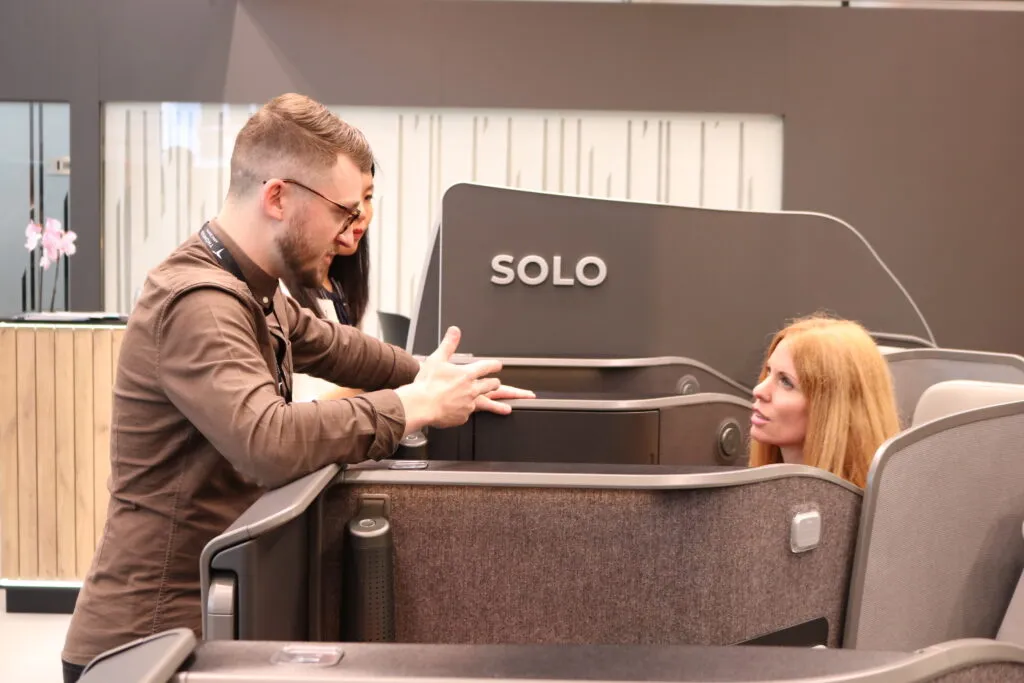 A Thompson executive is speaking to Mary Kirby as she is testing out the VantageSOLO seat on the show floor at AIX. SOLO can be installed on the A220.