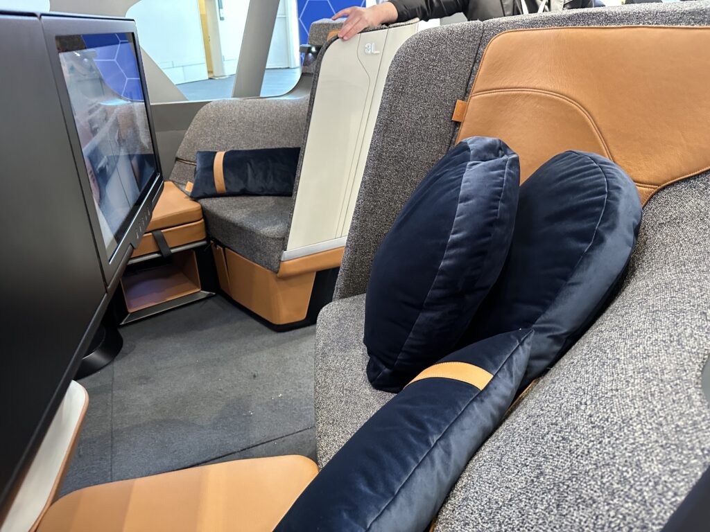 Side view of the Optimares SoFab business class seat in speckled grey with light brown details and blue pillows. It also includes a large IFE screen.