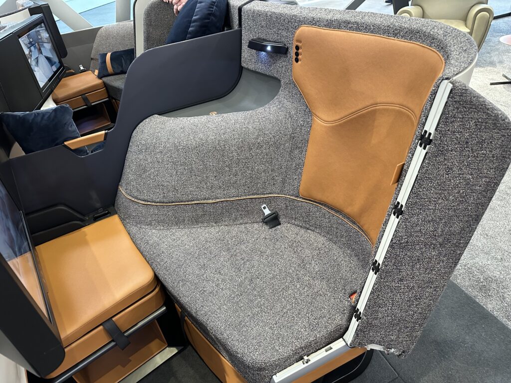 Optimares soFab business class seat is in all speckled grey with light brown details in the seatback and a blue pillow. It also includes a large IFE screen. Cabin interiors