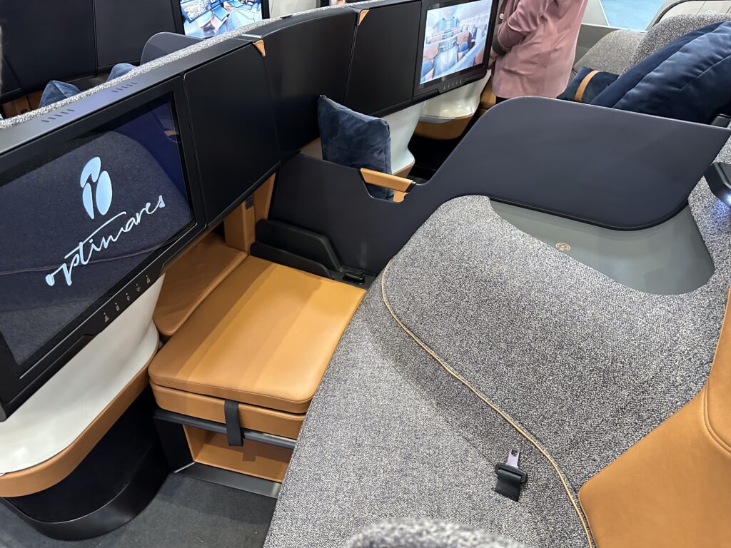 Optimares SoFab business class seat is in speckled grey with light brown details in the seatback and a blue pillow. It also includes a large IFE screen. The only moving parts are a pull-out cushioned storage area.