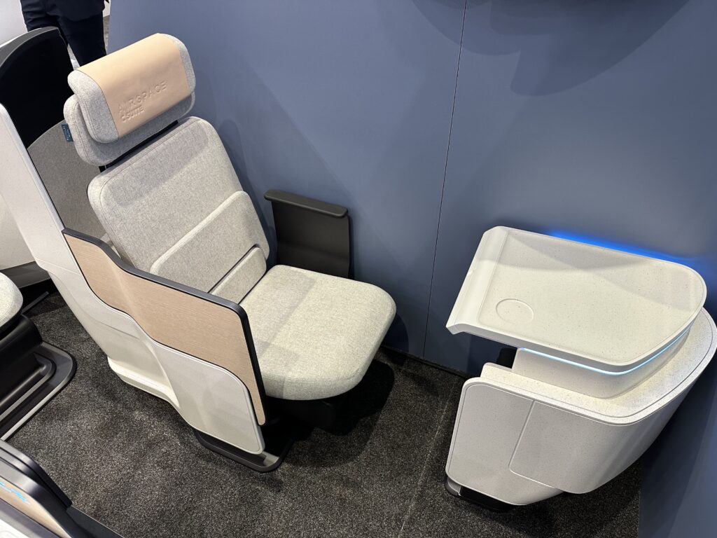 A display concept model of the C Suite by Airbus and Stelia. This is a business class seat in mostly grey fabrics with a beige headrest cover and sidewall detailing. The armrest of the window seat, pictured, is black and rather petite. 