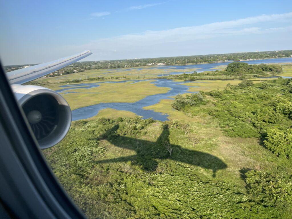 An aircraft shadow is seen on the green landscape below, as the aircraft is flying over it. This image was shot through the window of the seated passenger.