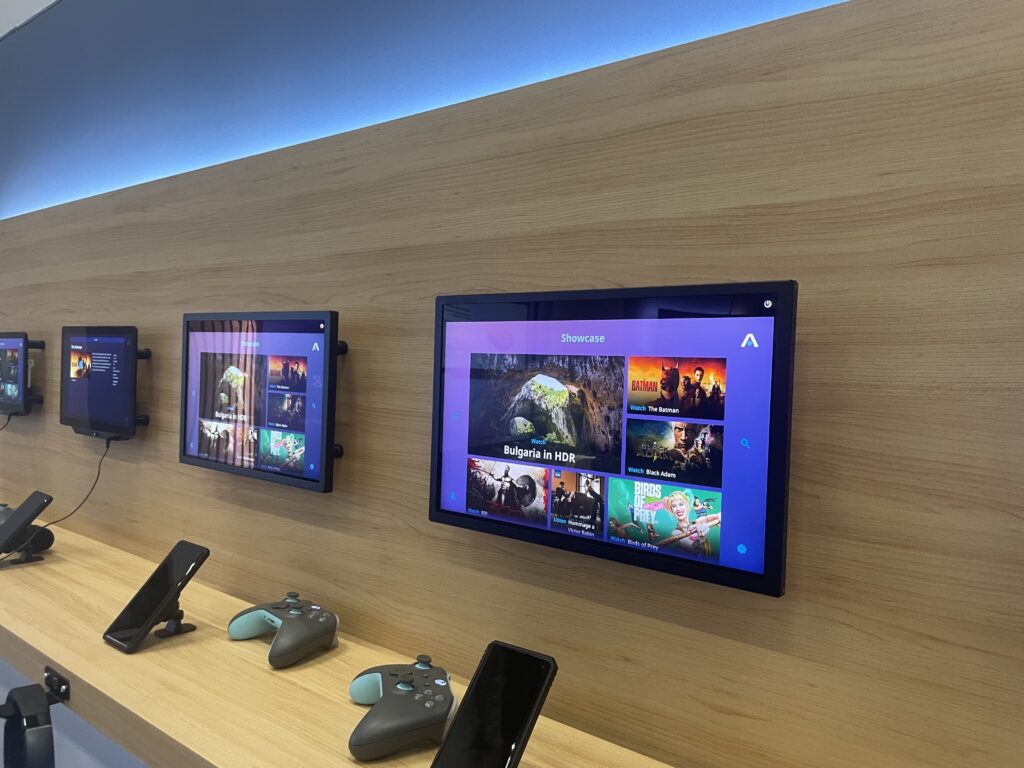 Three Thales AVANT Up IFE screens are displayed on a wall, whilst PEDs and gaming controllers sit below them on a shelf.