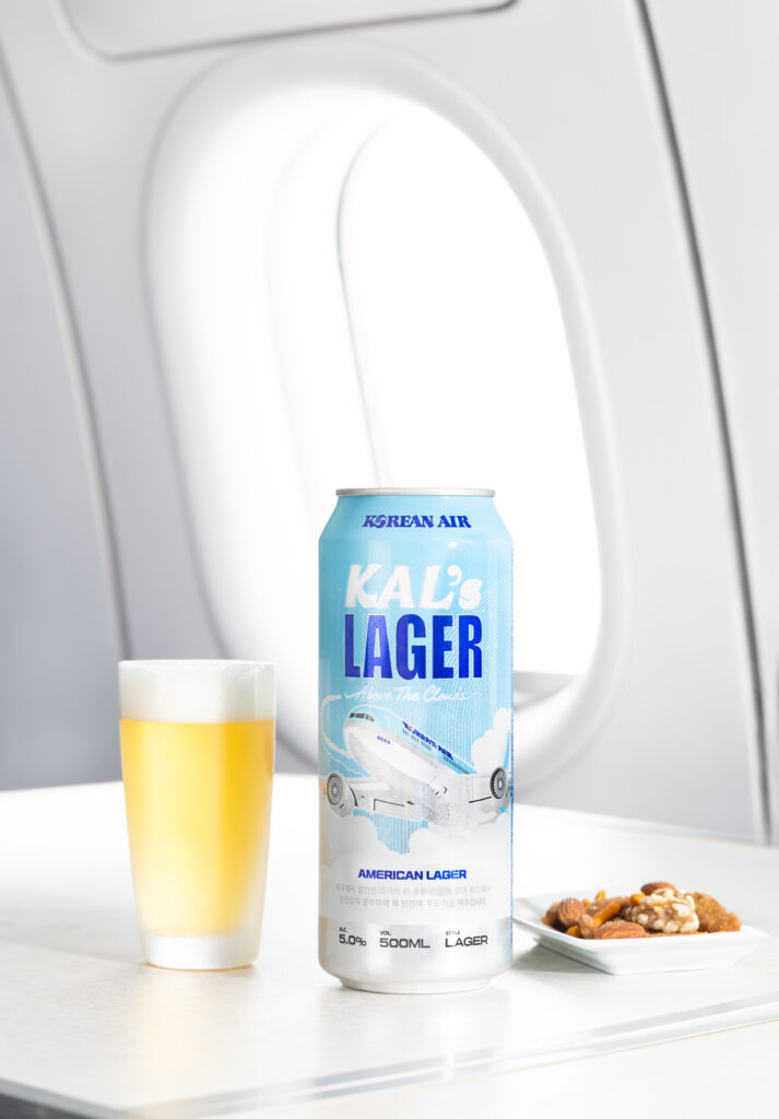 A tall can of KALs Lager is sitting next to a full glass of beer and a handful of nuts.