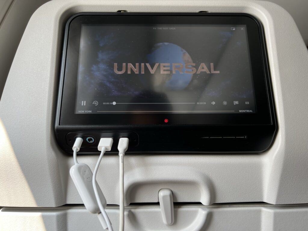 The Universal logo is displayed on the seatback IFE. Devices are plugged into its power and sound outlets. 