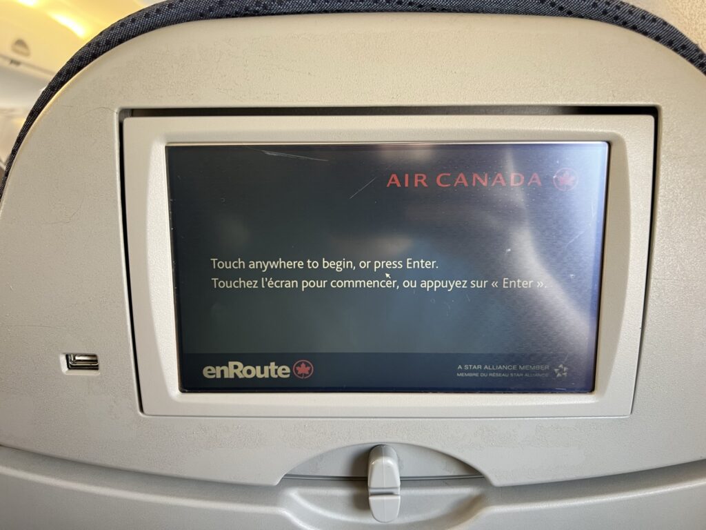 The seatback IFE screen on board the Air Canada E175. The screen says: "Air Canada and "touch anywhere to begin or press enter".