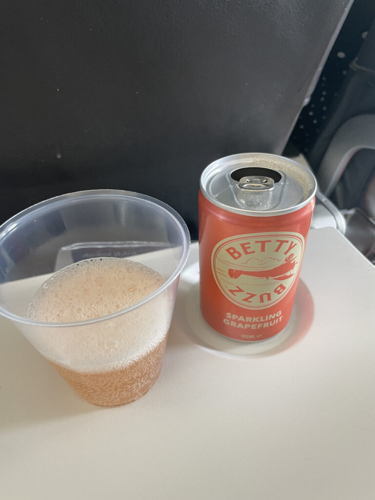 Betty Buzz soda in an orange can sits alongside a plastic cup filled with the drink on the BA A319 tray table.