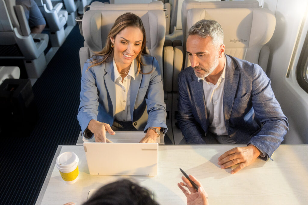 Male and female business class passengers, sitting side by side, at a table, whilst the woman uses her laptop. 