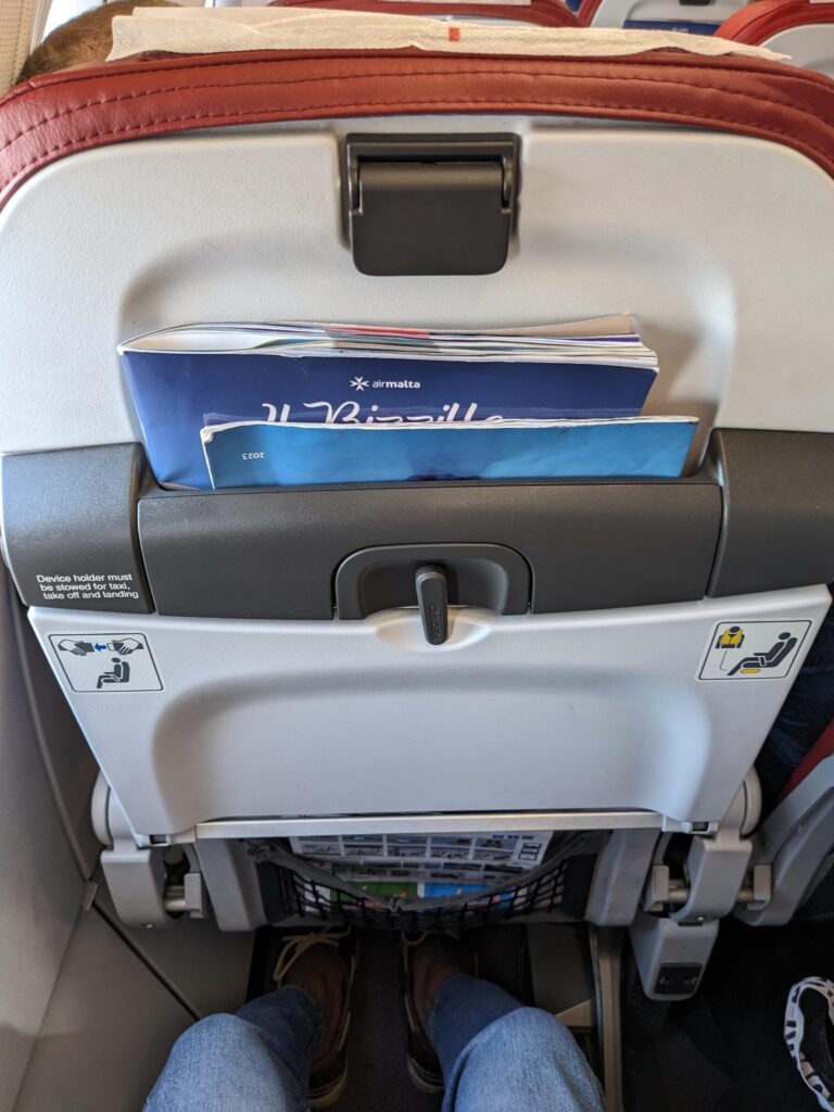 The back of an Air Malta aircraft seat shows a literature pocket, stowed tray table and a red seat cover. The passenger's legs clearly have room to move.