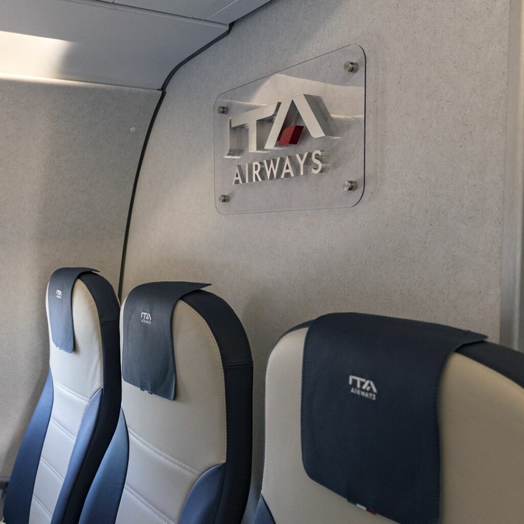 ITA Airways crest by ABC International is seen above three economy class seats. Each blue headrest also displays the ITA logo.