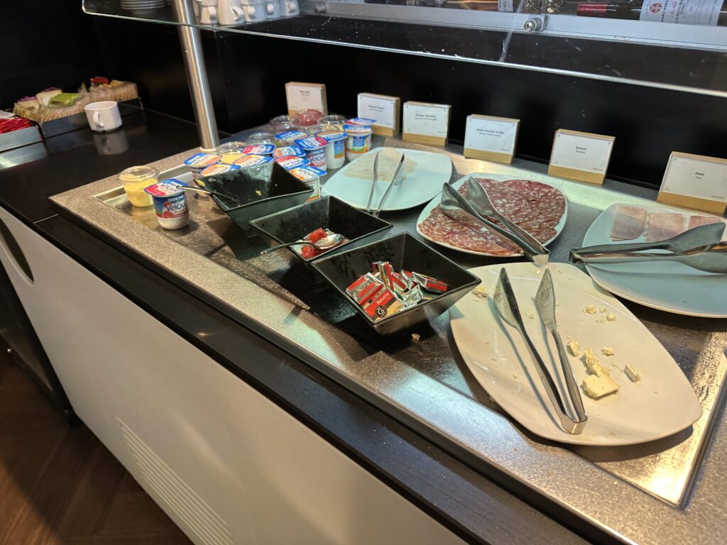 A picked over cold section of the buffet with some assorted meats, yogurt cups and an almost empty plate of cheese. 