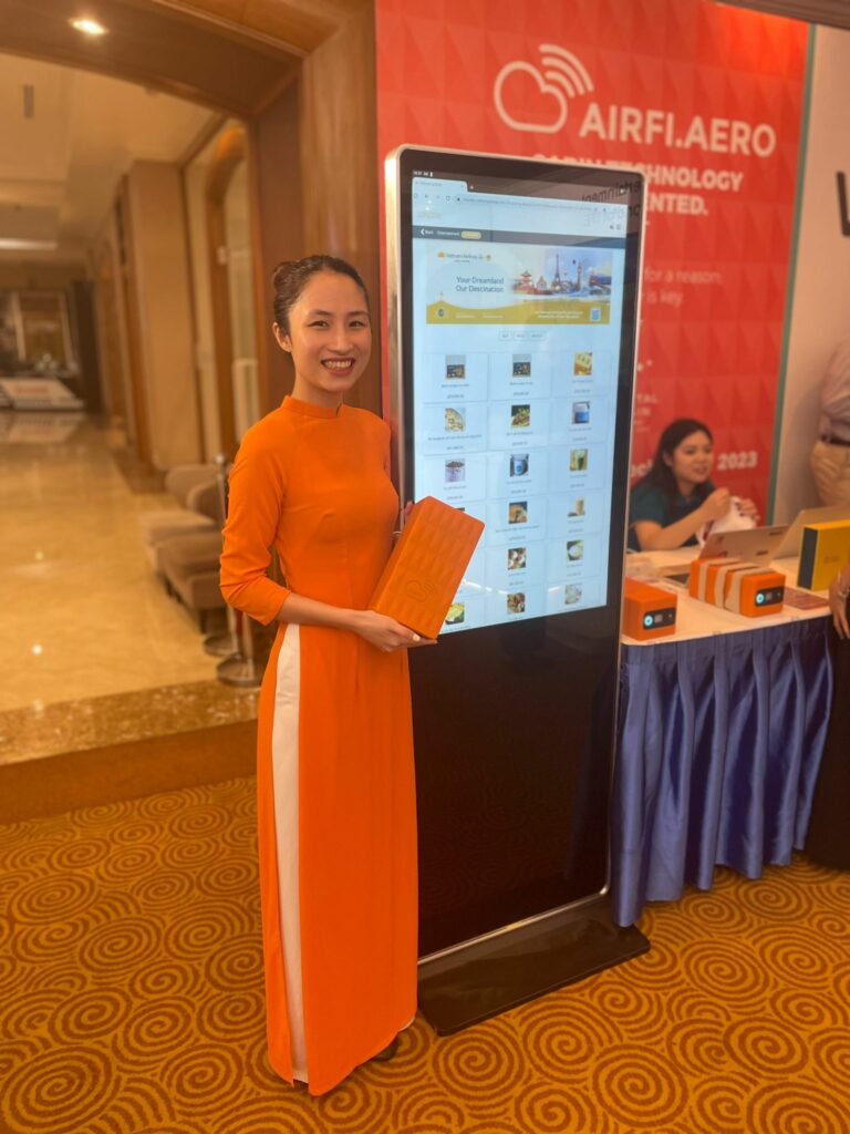 A female Vietnam Airlines representative is holding an AirFi box in front of a large mock mobile device displaying AirFi.