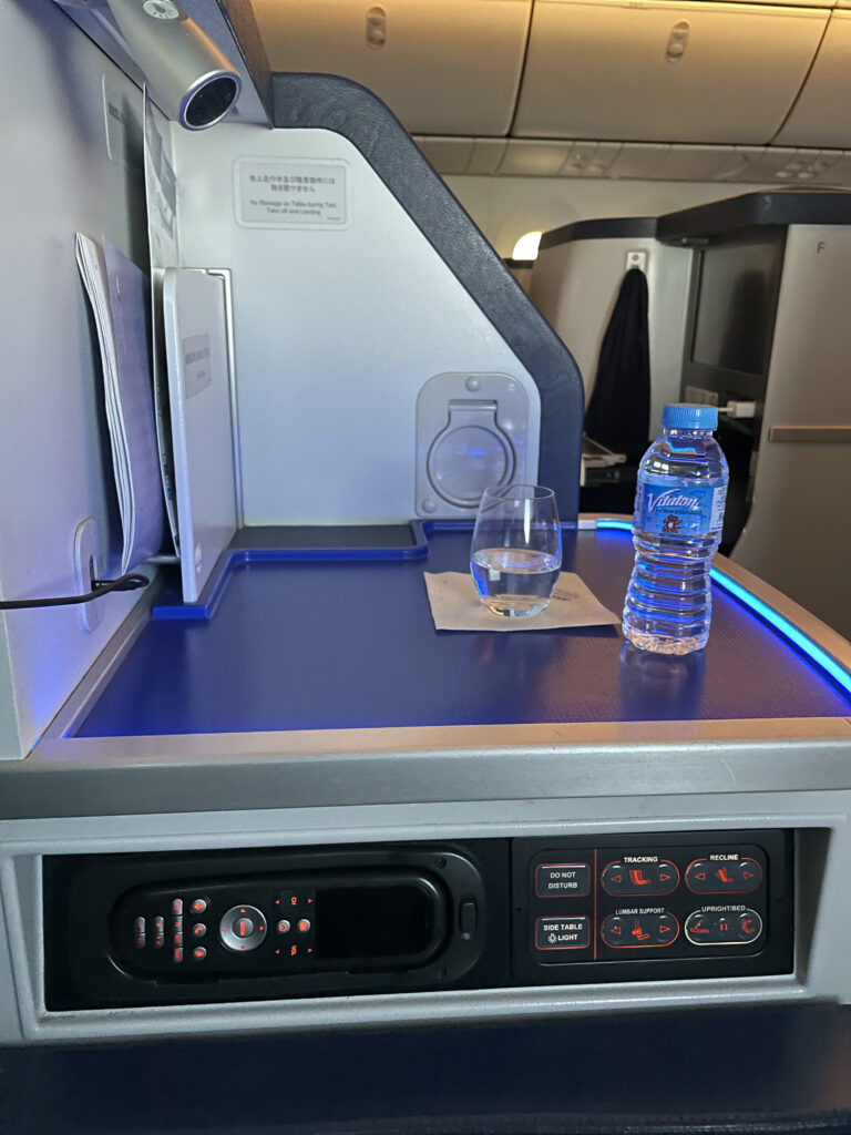 An assortment of seat controls at the side of the ANA business class seat table. A bottle of water sits atop the side table of the suite.