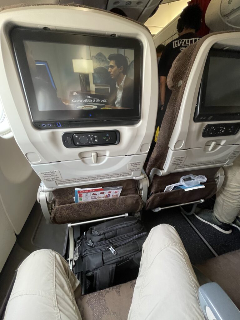 A man's knees are shown in a decent proximity from the aircraft seat, with seatback IFE, directly in front of him. 