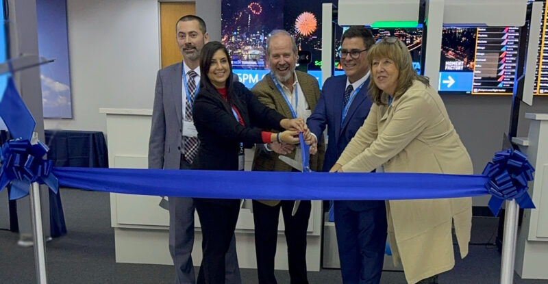 Pictured at the NTO pre-production center ribbon cutting ceremony, left to right: Dave Rellinger, Vice President, US Branches, Schneider Electric; Luna Katbah, Director Airport Systems, NTO; Jim Durkin, Executive Vice President, Tishman Construction; Anthony Natale, Sales Vice President North America, SITA; Faith Varwig, Managing Principal, Faith Group. SITA ribbon cutting at JFK. 4 people are ceremoniously cutting a blue ribbon.