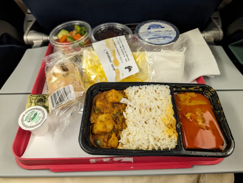 A meal of rice, veggies and meat is served with other snacks and displayed on the Air India 777 economy class tray table.