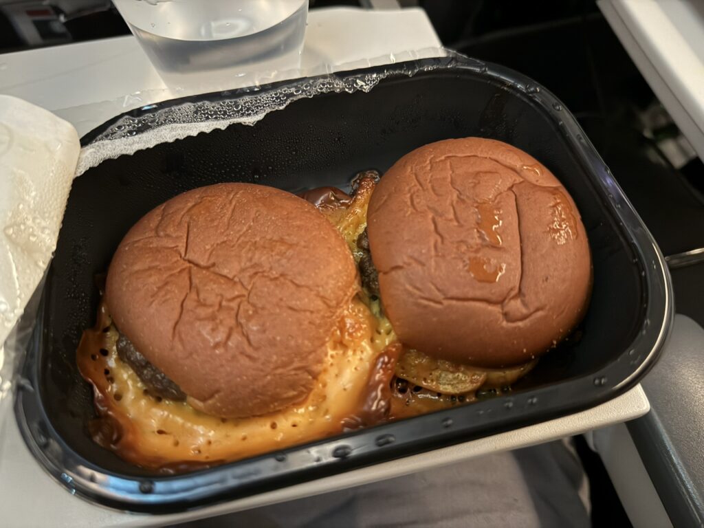 Two cheeseburger sliders in a plastic container. The over-melted cheese is messy.