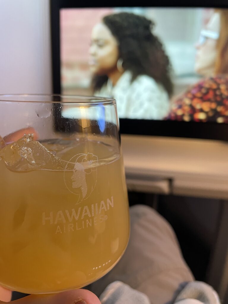 A passenger is holding up a drink in front of the IFE screen with a woman on the screen. 