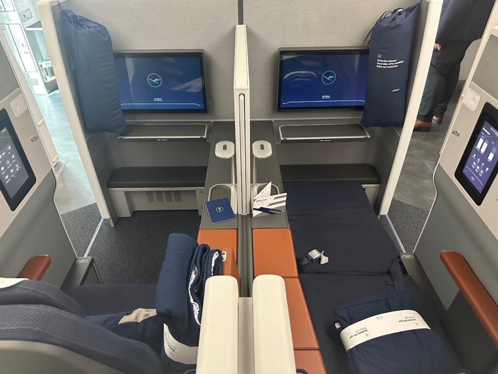 Picutred: Lufthansa Allegris front row, biz-plus middle pair enabling a more social experience if chosen. Blues and beiges color the scene of two suites, one in bed mode, and large IFE screens.