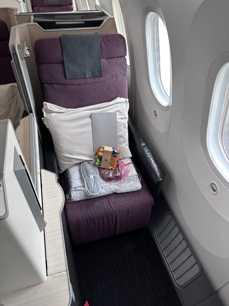 JAL business class 787 Collins Apex seat. The seat is a burgundy with grey detailing. A blanket, pillow, slippers and amenity kit sit on the seat.