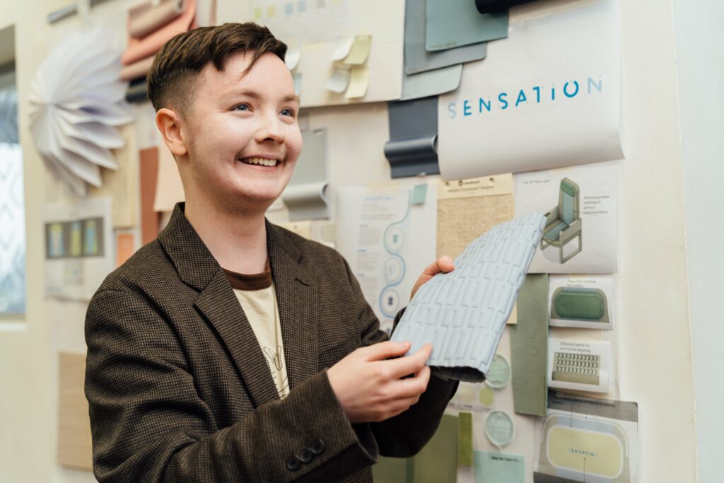 O'Donnell showcases the material for the Murihead seat covers. This swatch happens to be a light blue color. Various details and swatches of the collection are on the wall behind O'Donnell.