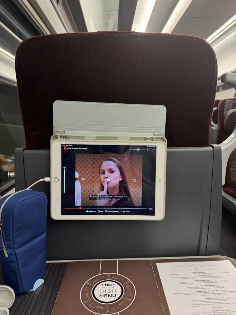 An iPad is lodged in the seat surround of the seat in front of the author. It is playing a movie.