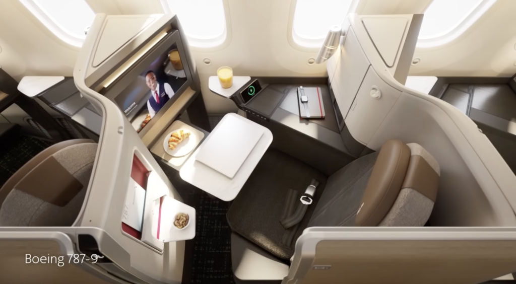 Overhead view of a window seat in American Airlines business class. Light shines in through the window onto browns and greys. A meal is set out for the passenger to enjoy.