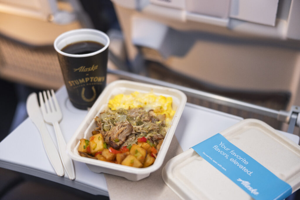 Carnitas Breakfast Bowl is pictured on a tray table with a cup of coffee.