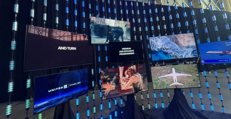 Lots of large IFE displays hang on a wall, showcasing different channels, including in the bottom left, a screen that stays "Starlink/United" to represent the integration of IFC and IFE.