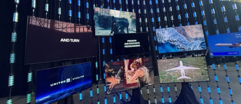 Lots of large IFE displays hang on a wall, showcasing different channels, including in the bottom left, a screen that stays "Starlink/United" to represent the integration of IFC and IFE.