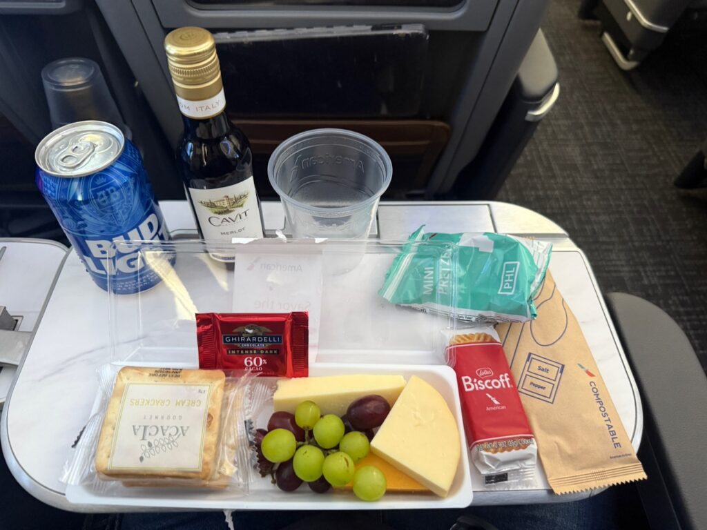 Cheese, fruit and a bottle of Cavit wine sit beside a can of Bud Light and some Biscoff cookies on the tray table.