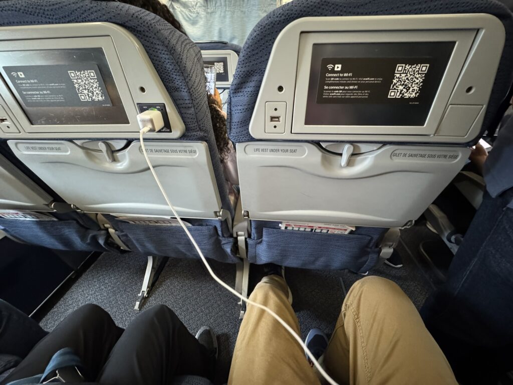 The Air Canada seatback screens on the A320 are covered with a sticker featuring a QR code that directs passengers to the free Wi-Fi. A personal device is plugged into the AC outlet. 