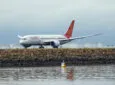 An Air India Boeing B787-8 plane, registration VT-ANZ, taking off to the north from Sydney Kingsford-Smith Airport and heading to Delhi as flight AI301. In the background are fuel storage tanks. A buoy on the water marks the airport exclusion zone. This image was taken from near Kyeemagh Beach, Botany Bay on an overcast and rainy morning on 17 March 2024.