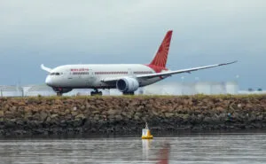 An Air India Boeing B787-8 plane, registration VT-ANZ, taking off to the north from Sydney Kingsford-Smith Airport and heading to Delhi as flight AI301. In the background are fuel storage tanks. A buoy on the water marks the airport exclusion zone. This image was taken from near Kyeemagh Beach, Botany Bay on an overcast and rainy morning on 17 March 2024.