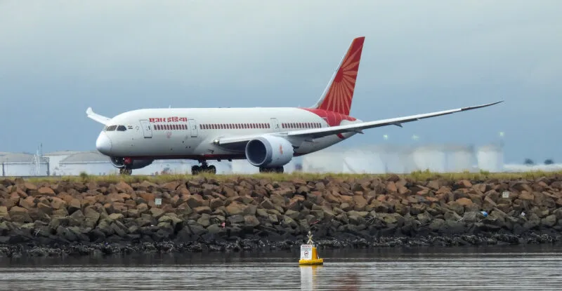 An Air India Boeing B787-8 plane, registration VT-ANZ, taking off to the north from Sydney Kingsford-Smith Airport and heading to Delhi as flight AI301. In the background are fuel storage tanks. A buoy on the water marks the airport exclusion zone. This image was taken from near Kyeemagh Beach, Botany Bay on an overcast and rainy morning on 17 March 2024.