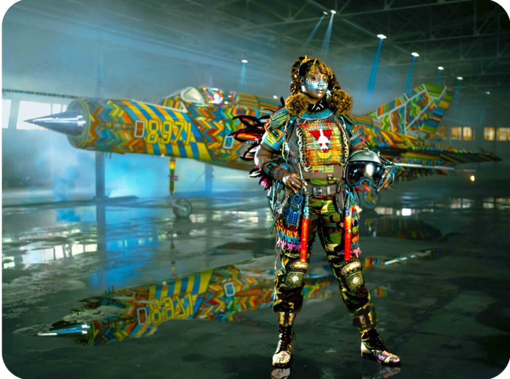 A MiG-21 military flight suit is being modeled by a woman of African descent. She is covered in a brightly colored tribal-inspired armor featuring colorful beads.